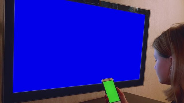 Teenage girl sits in front of TV set holding smartphone