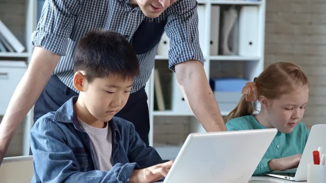 Young Male Teacher Standing Close To His Two Primary School Students And Monitoring Their Progress While They Are Doing Task On Laptop Computer