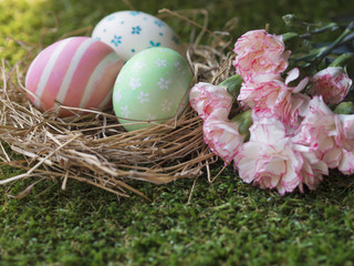 Sweet pink, green and white painting Easter eggs in a bird nest with beautiful spring flowers, carnation on green grass in spring garden