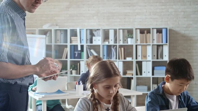 Students of primary school sitting in computer coding class and doing task on laptops while their teacher is using tablet and monitoring their progress
