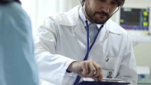 Medium shot with tilt down of bearded male doctor in lab coat listening to breathing of female patient with stethoscope, then writing notes on clipboard