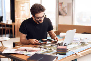 Freelancer bearded man looking at color swatches at laptop sitting at desk.