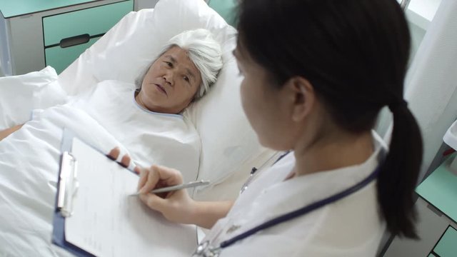 High angle shot of recovering senior woman lying in hospital bed and talking with caring female doctor making notes and supporting her