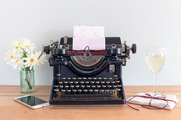Antique typewriter with note card in the carriage, on a wooden table surrounded by flowers, glass of wine, phone and a bundle of notes tied with a string. Concepts of home office, thank you, nostalgia