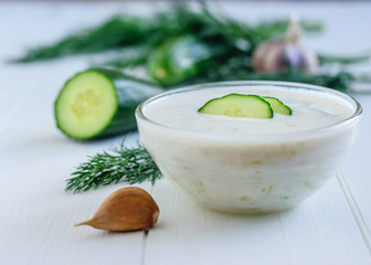 Freshly made tzatziki in a glass bowl with cucumber on a white wooden table.