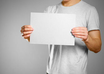 A man in a gray t-shirt keeps a grey clean blank sheet of A4. Isolated on grey background