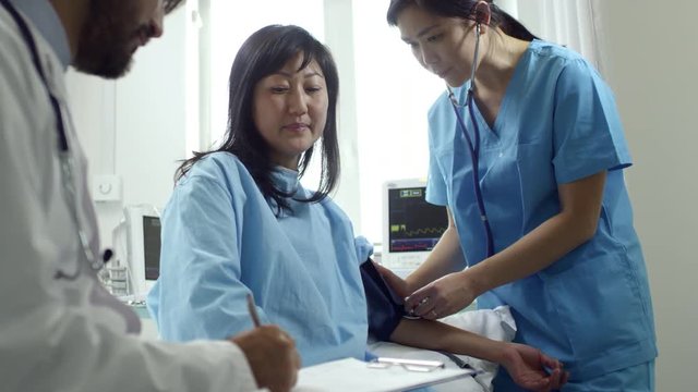 Medium shot of unrecognizable male doctor in lab coat talking with female patient as young nurse in scrubs measuring her arterial blood pressure