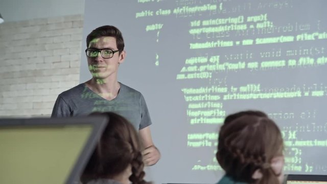 Young male teacher of information technology explaining new topic to primary school students with the help of projector screen, then two girls raise hands