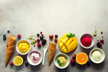 Top view red, pink, yellow, green, white ice cream balls in bowls, waffle cones, berries, orange, mango, pistachio, grey concrete background. Ice-cream colorful collection, flat lay, summer concept