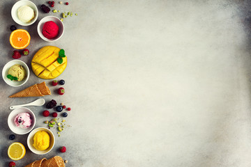 Top view red, pink, yellow, green, white ice cream balls in bowls, waffle cones, berries, orange, mango, pistachio, grey concrete background. Ice-cream colorful collection, flat lay, summer concept