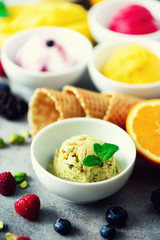Red, pink, yellow, green, white ice cream balls in bowls, waffle cones, berries, orange, mango, lemon, mint, pistachio, grey concrete background. Ice-cream colorful collection, summer concept