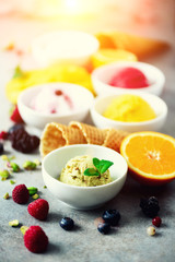 Red, pink, yellow, green, white ice cream balls in bowls, waffle cones, berries, orange, mango, lemon, mint, pistachio, grey concrete background. Ice-cream colorful collection, summer concept