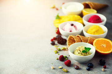 Red, pink, yellow, green, white ice cream balls in bowls, waffle cones, berries, orange, mango, lemon, mint, pistachio, grey concrete background. Ice-cream colorful collection, summer concept