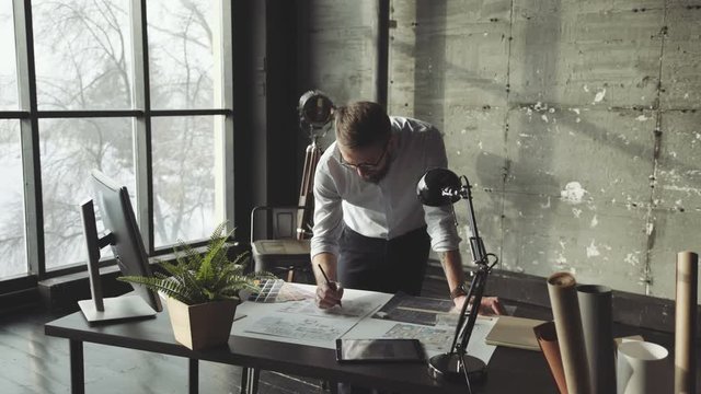 A spacious light office in loft style with panoramic windows. A male businessman is working, standing in front of the table, making notes on the blueprints. A modern architect is working on a project.