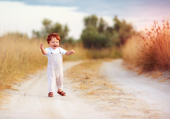 adorable redhead toddler baby boy in jumpsuit running along rural summer road in sunburned field