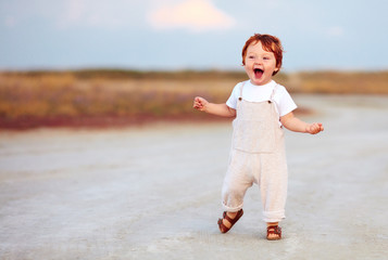 adorable redhead toddler baby boy in jumpsuit running through the summer road and field
