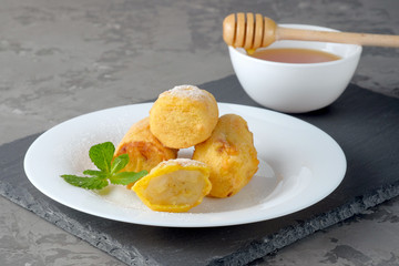Bananas in batter and sprinkled with powdered sugar on a white plate on the gray slate background. The bowl with honey near it.