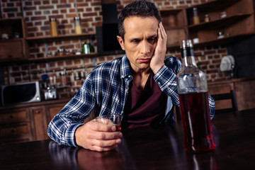 upset man sitting at table with bottle and glass of alcohol at home