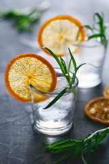 Two shots of cocktails with ice, dried orange slice and fresh tarragon on a grey table.