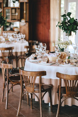 Decorated wedding table in rustic style for dinner with white and beige tablecloths, wine glasses for flowers and Viennese wooden chairs