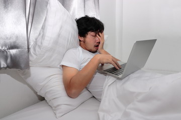 Tired stressed young Asian man feeling sleepy while using laptop on the bed in bedroom.  Hard work concept.