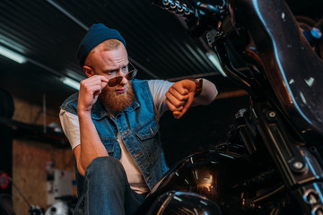Fototapeta na wymiar handsome young man in sunglasses looking at watch while sitting on motorcycle at garage