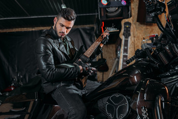 handsome young man playing guitar while sitting on bike at garage