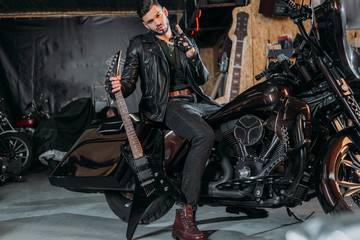 Fototapeta na wymiar stylish young man in leather jacket sitting on bike with electric guitar at garage