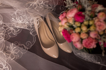 Beige bridal shoes and a veil lying on a red armchair. Wedding bouquet with purple and pink roses out of focus.