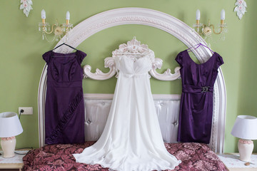 White wedding dress and two lilac bridesmaid’s dresses on a hanger on the wall. Morning of the bride. Fees. Wedding