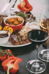 plate with sauces, roasted potatoes, grilled meat with vegetables and glasses of wine on table