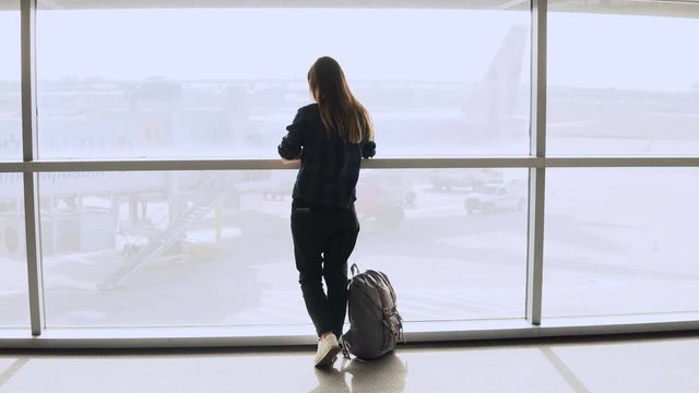 Young girl takes selfie photo near airport window. Happy European tourist with backpack uses smartphone in terminal. 4K.