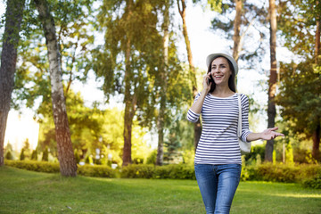 An attractive smiling girl in casual clothes speaks on the phone strolling in the park.