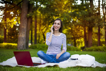 How are you? Attractive Young modern woman in casual clothes talking on the phone with her sister sitting on a plaid in a city park.