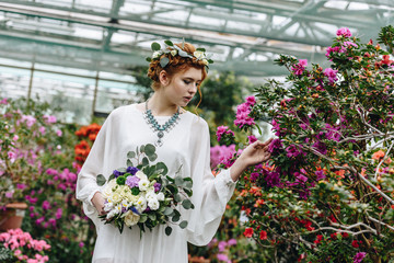 beautiful elegant young bride holding wedding bouquet and touching flowers in botanical garden