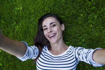 An attractive smiling modern young woman with chic hair and in casual clothes lies on the green grass. View from above