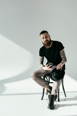 handsome bearded tattooed man sitting on chair with smartphone and hat on white