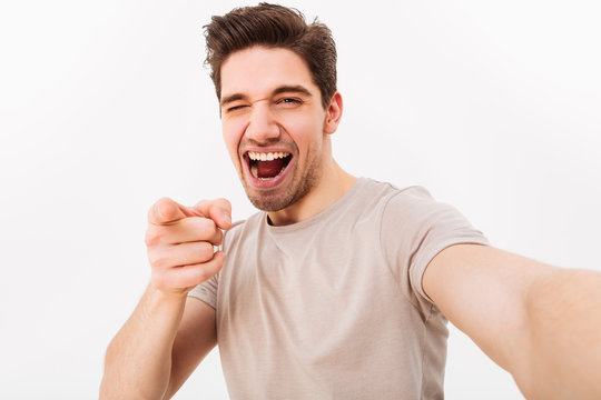 Joyful man with brown hair gesturing index finger on camera meaning hey you while taking selfie, isolated over white background
