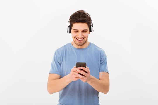 Attractive man with short dark hair listening to music via wireless headphones and holding cell phone, isolated over white background