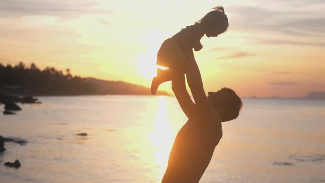 Happy father and daughter playing on the beach at amazing sunset with lens flare effects. Dad holds cute girl in hands. slow motion. 3840x2160