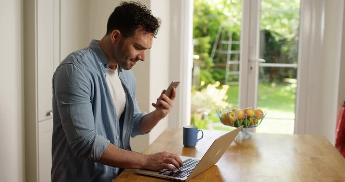 4K Happy casual man using laptop at home & having flirtatious phone conversation. Slow motion.
