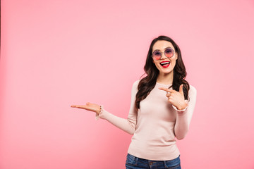 Modern caucasian woman in casual pointing finger on advertising product or text while holding copy space on her palm, isolated over pink background
