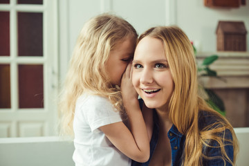 I'll tell you a secret. Cheerful smiling beautiful blond mother and daughter embrace sitting on the couch at home. Mothers Day. Women's Day. March 8.