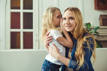 I'll tell you a secret. Cheerful smiling beautiful blond mother and daughter embrace sitting on the couch at home. Mothers Day. Women's Day. March 8.