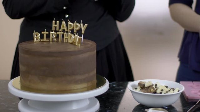 Cake with sign Happy birthday is on the table near two confectioners. Professionals are standing at the desk with small bowl with white and black chocolate. Woman in gloves is taking a piece to