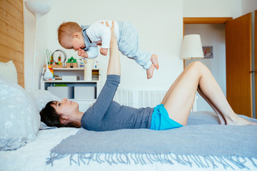 Side view portrait of middle age white caucasian mother and infant baby boy together on bed in bedroom doing physical fitness exercises yoga together, early development.