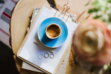 Delicious freshly brewed morning espresso coffee a notepad and a book on the rustic wooden slab table with some flowers blurred on the foreground, flat lay