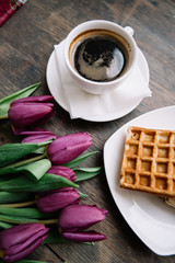 Delicious fresh morning hot cup of black coffee with waffles and purple tulip flowers on the rustic wooden table background