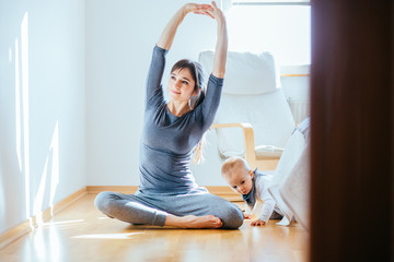 Cute european mother doing yoga or pilates while her infant baby boy playing near at cozy simple home interior. Motherhood, healthy lifestyle and people concept.
