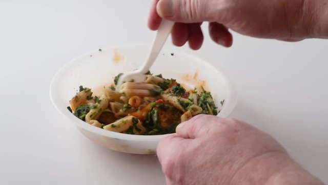 Video of a cooked meatball, pasta and spinach in a marinara sauce TV dinner in a plastic tray being stirred with a fork on a white counter top.